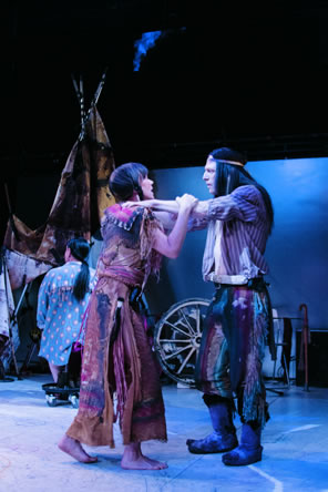 Troilus with long black hair and a headband, wearing striped, open shirt, multicolor pants to his shins and animal skin boots has his arms on Cressida's shoulders as she grasps his arms: she is in multi-color, doublslitted buckskin dress with tassles, pigtails and bare feat. In the background is a teepee, wagon wheel, and Pandorus in a polka dot blue shirt and long pony tail, his back to us.