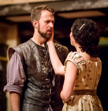 Leantio is wearing a leather renaissance vest with buckles down the front and a purple shirt with rolled up sleeves. Bianca in simple tan dress with a pattern of brown brances and leaves holds her right hand on Leantio's bearded cheek.