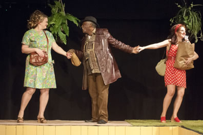 Falstaff in brown leather jacket and brown pants and shirt and hat holding a bagged liquor bottle holds the hands of Mistress Ford in green-patterned knee-length dress and brown purse, and Mistress page in white polka dot red thigh-length dress, holding a bag of groceries, with hanging planters of ferns behind.