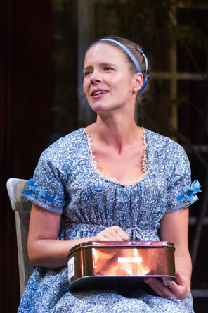 Marianne wearing her simple blue print dress with lace trim and blue ribbons tied in bows on each sleeve, sits on a chair, a tin box in her hands,