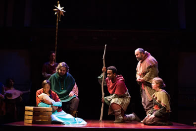Mary ina long blue dress sits next to a crate and cradles in infant in her arms, an orange shawl draped over her shoulder, Josephe in green cloak sits on a crate behind her, and across from them, Coll with his staff kneels on one knee, Gib stands with head bowed, and Daw kneels on both knees. An actor holds the gold Star of Bethlehem ona pole in the background, and Kay plays a lute.