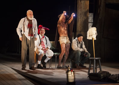 Lear stands in white untucked shirt, brown pants, suspenders, and red and brown scarf about his kneck, fool in white knee pants, socks, shirt, black suspenders, red-striped tie, red nose and white face with coxcomb sits, Edgar as Poor Tom with blanked around him and only in white cutoffs stands holding up two flashlights, and Kent in black stocking cap, gray hoody, fingerless gloves, and blue jean sits, all ecept the Fool looking at a mob stuck into a joint stool; Fool is looking up at Lear.