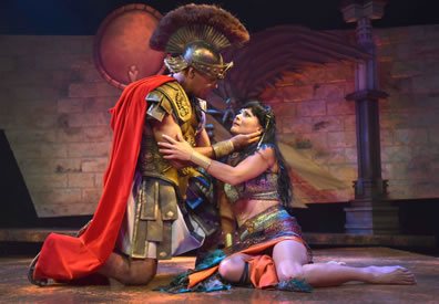 Antony in classic Roman legion uniform, skirt, leather armo, crest helmet and red ape on his knees stroking Cleopatra's neck as she holds his arm, she sitting on the floor in a multi-colored sheen dress, halter, and bare midriff
