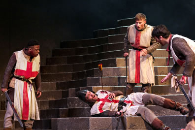Three soldiers wearing Saint George smocks over chain-mail armor stand around another soldier lying on the stone steps with an arrow sticking into his shoulder, blood on the smock, his hand gripping at his breast, a sword in his other hand.