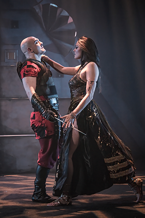 Antony in black armor over a red tunic and pants with black splots, black long gloves and boots, holding a sword as Cleopatra in a glittering black and gold dress showing her full leg as she strides to Antony, her right hand on his shoulder and her left resting on the blade of his sword. A portion of the stone pyramid is in the background, and the floor is lit in a tapestry of black and brown.
